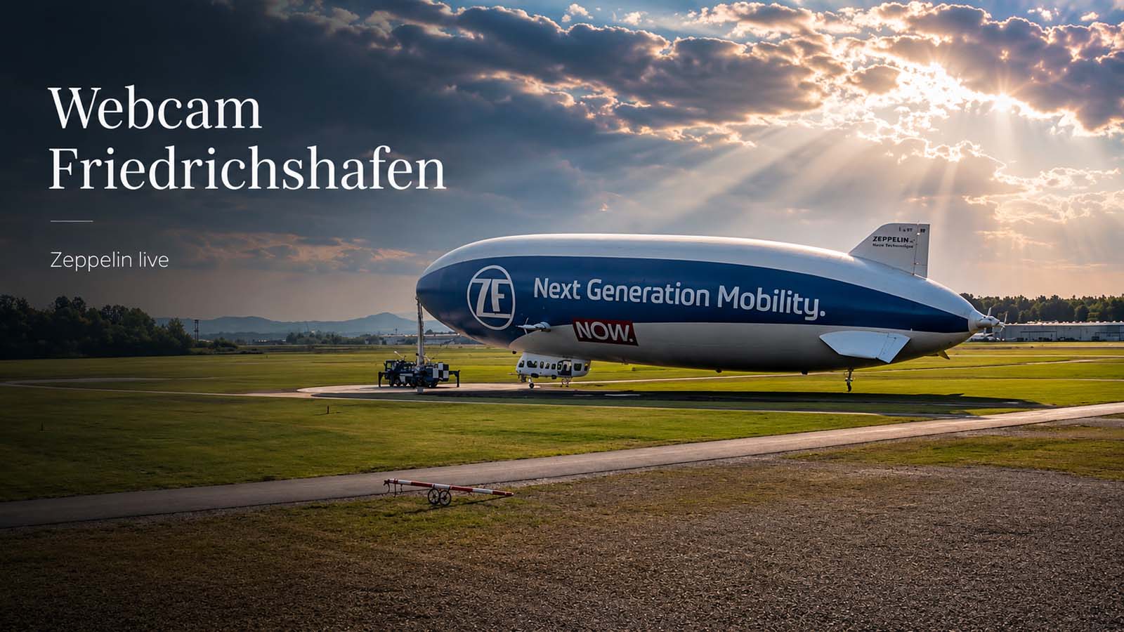 Zeppelin NT auf dem Flugfeld in Friedrichshafen unter einem dramatischen Himmel mit dem Text „Webcam Friedrichshafen – Zeppelin live“.